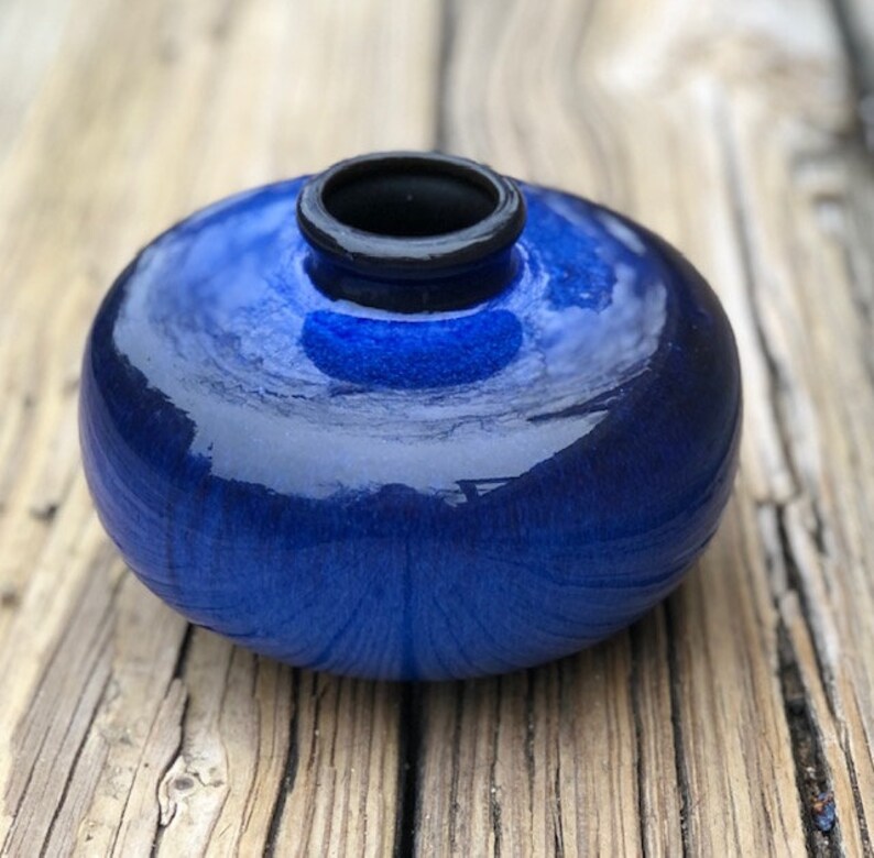 May include: A small, round, blue ceramic vase with a black rim. The vase has a glossy finish and is sitting on a wooden surface.