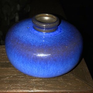 May include: A small, round, blue ceramic vase with a dark blue glaze. The vase has a wide mouth and a narrow neck.