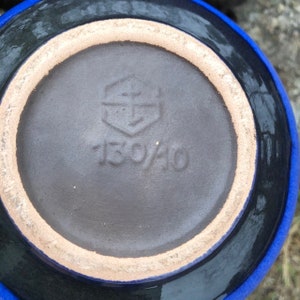 May include: Close-up of the bottom of a dark blue ceramic vase with a brown glaze. The vase has a hexagonal mark with an anchor inside and the number "150/40" printed on the bottom.