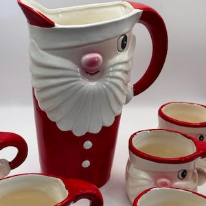 Può includere: Un set composto da una brocca e tazze in ceramica a forma di Babbo Natale. La brocca presenta una barba bianca, un naso rosa e un abito rosso con bottoni bianchi. Le tazze sono modellate come volti di Babbo Natale con bordi e manici rossi.