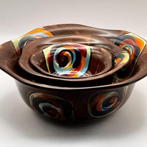 May include: A set of four brown ceramic nesting bowls. Each bowl features a unique, colorful spiral design in shades of red, orange, blue, and yellow. The bowls have wavy edges.