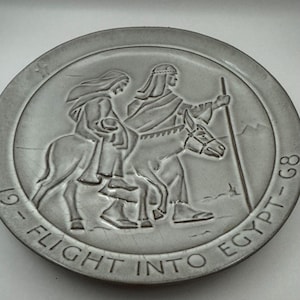 Puede incluir: Un plato de cerámica blanco con un diseño en relieve de la Huida a Egipto. El plato tiene el texto "19 - FLIGHT INTO EGYPT - 68" alrededor del borde.
