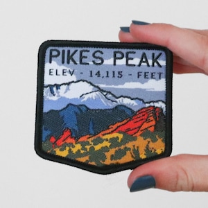 May include: A black and white embroidered patch with a colorful image of Pikes Peak, a mountain range with snow-capped peaks. The text "PIKES PEAK" is in black lettering above the image, and the text "ELEV - 14,115 - FEET" is in black lettering below the image.