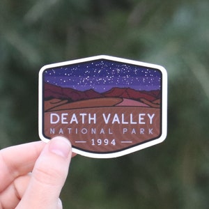 May include: A sticker featuring a landscape of Death Valley National Park under a starry night sky. The sticker has a white border and includes the text "DEATH VALLEY NATIONAL PARK - 1994". The background is a blurred green.