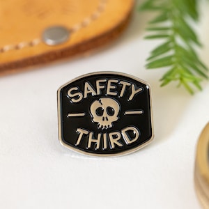 May include: A black and silver enamel pin with a skull and crossbones and the text "Safety Third".