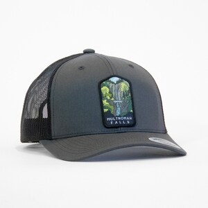 May include: A gray and black trucker hat with a patch featuring a depiction of Multnomah Falls. The patch has the text "Multnomah Falls" on it.