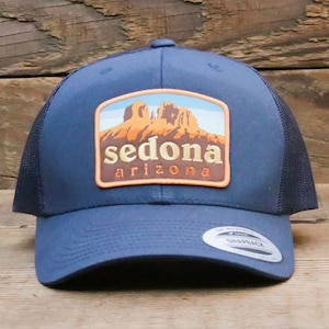 Peut inclure: Casquette de camionneur bleu marine avec un dos en filet et une visière incurvée. L'avant présente un écusson rectangulaire avec le mot "sedona" au-dessus de "arizona" et une image de formations rocheuses rouges. Fermeture à pression.