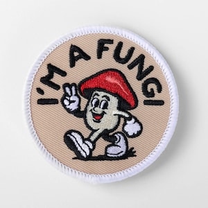 May include: A circular embroidered patch with a beige background and a white stitched border. It features a cartoon mushroom character with a red cap, white gloves, and shoes, making a peace sign. The text I'M A FUNGI is arched above the character.