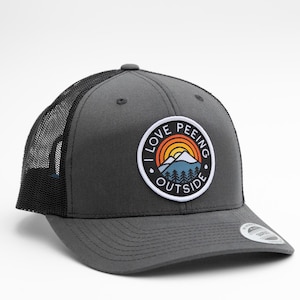 May include: A charcoal gray trucker hat with a black mesh back. The hat features a circular patch with the text "I LOVE PEEING OUTSIDE" around a mountain and rainbow design. The hat has a curved brim.