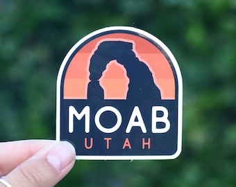 May include: A black and orange sticker with a silhouette of Delicate Arch, a natural sandstone arch in Arches National Park, Utah. The sticker says "Moab, Utah".