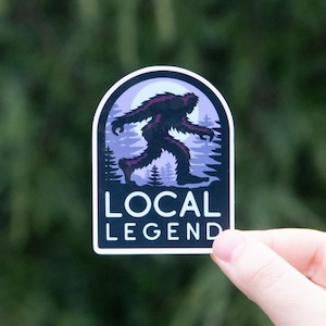 May include: A black and purple sticker with a silhouette of Bigfoot walking through a forest. The sticker has the text "LOCAL LEGEND" at the bottom.