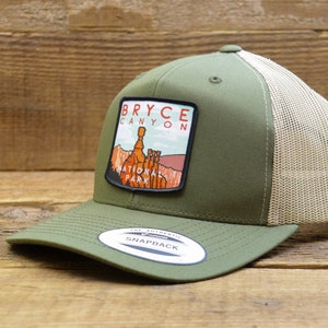May include: A green and beige trucker hat with a patch that says "Bryce Canyon National Park" and a picture of the park's iconic hoodoos. The hat has a mesh back and a snap closure.