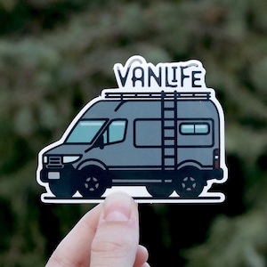 May include: A gray vanlife sticker with a black outline. The van has a ladder on the side and a roof rack. The text "VANLIFE" is printed on the roof rack.