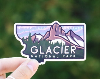 Könnte beinhalten: Ein Aufkleber mit einer stilisierten Illustration des Glacier National Park in Montana. Der Aufkleber hat die Form des Bundesstaates Montana und zeigt eine Bergkette mit einem rosa und violetten Sonnenuntergang im Hintergrund. Der Text "Glacier National Park" ist in Weiß unter der Illustration gedruckt.