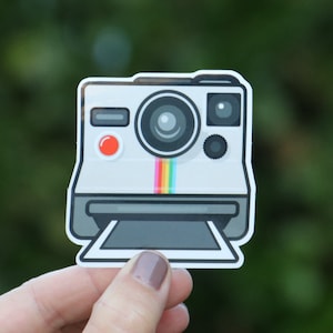 May include: A white and gray cartoon illustration of an instant camera with a rainbow stripe across the front. The camera has a red button and a black lens.