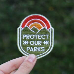Fingers pinch the lower edge of the Protect Our Parks patch with dense shrubbery behind it.
