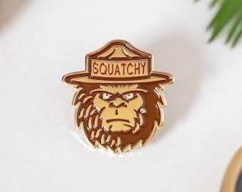 Puede incluir: Pin de esmalte de color dorado con un dibujo animado de un sasquatch marrón que lleva un sombrero de guardabosques con el texto "SQUATCHY" en el sombrero.