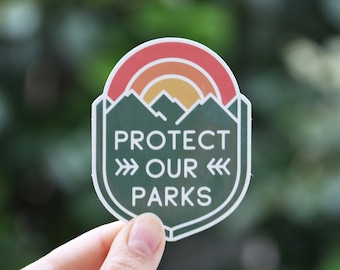 May include: A green and white sticker featuring a mountain range with a sunset behind it. The sticker reads "Protect Our Parks" with arrows pointing in opposite directions.