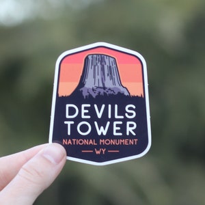 May include: A sticker featuring a stylized illustration of Devils Tower National Monument in Wyoming. The sticker is dark blue with a sunset orange background and white text.