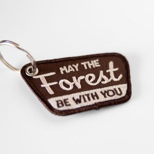 May include: A brown trapezoidal embroidered patch keychain with a silver metal ring. The patch features white embroidered text that reads MAY THE Forest BE WITH YOU, with the word Forest in a larger, stylized script font.