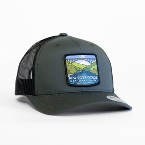 May include: A dark green trucker hat with a black mesh back. The hat features a patch with a colorful illustration of a bridge and the text "New River Gorge National Park 1983".
