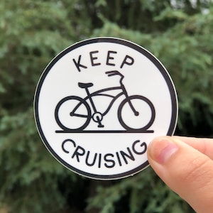 May include: A white round sticker with a black outline. The sticker features a bicycle with the text "KEEP CRUISING" around the bicycle.