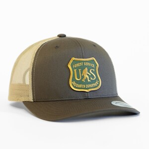 May include: A brown and tan trucker hat with a green and gold patch that says "Forest Service US Sasquatch Department".