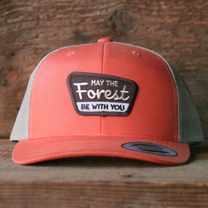 May include: A coral orange and gray trucker hat with a brown patch that reads "May the Forest Be With You".