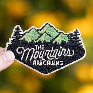 May include: Embroidered patch with a mountain and forest design. The patch is black with green mountains and trees, and the text "The Mountains Are Calling" in white script. The patch is held against a blurred yellow and green background.