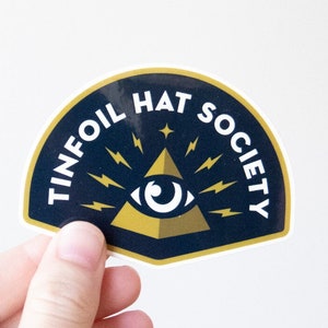 A hand holds the Tinfoil Hat Society sticker against a plain white wall, its gold eye and pyramid showing.