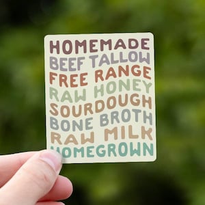 May include: A rectangular sticker with rounded corners featuring wavy, stacked text in shades of brown, green, and cream. The text reads: HOMEMADE, BEEF TALLOW, FREE RANGE, RAW HONEY, SOURDOUGH, BONE BROTH, RAW MILK, and HOMEGROWN in a retro font.