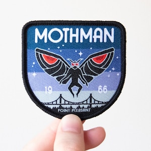 May include: A black and blue embroidered patch with the text "MOTHMAN" in white. The patch features a black moth with red eyes and a bridge in the background. The text "1966" is below the moth and the text "POINT PLEASANT" is below the bridge.