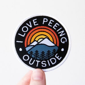 May include: A black and white round patch with a white border. The patch features a colorful mountain scene with a sunset and the text "I LOVE PEEING OUTSIDE".