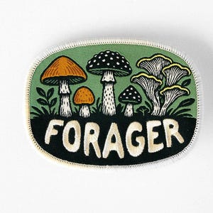 May include: An oval embroidered patch featuring a green background with various mushrooms in orange, white, and black. The bottom section is black with the word FORAGER stitched in large, cream-coloured capital letters. The patch has a white border.