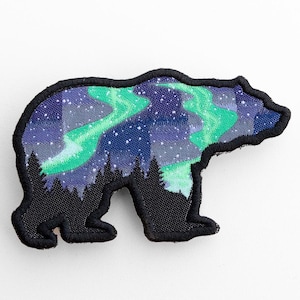Peut inclure: Écusson brodé noir en forme d'ours. L'intérieur de l'ours représente des aurores boréales, avec des tourbillons verts et bleus sur un ciel étoilé. Le patch est bordé de noir.