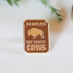 Pu&ograve; includere: Uno spillo in smalto marrone con finiture dorate che presenta una silhouette di bisonte e il testo "Do Not Pet The Fluffy Cows".
