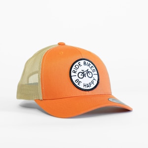 May include: Orange and tan trucker hat with a black and white patch that says "Ride Bikes Be Happy" and a bicycle graphic.