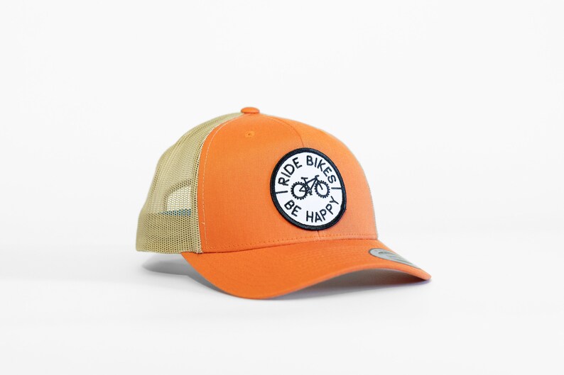 May include: Orange and tan trucker hat with a black and white patch that says "Ride Bikes Be Happy" and a bicycle graphic.