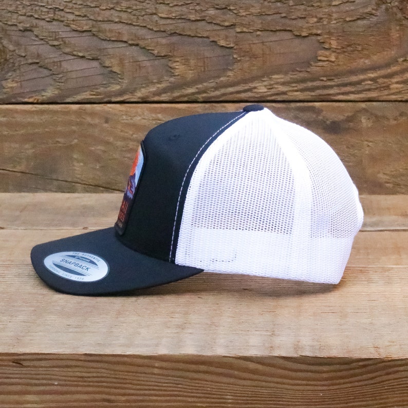 Op de afbeelding: Een zwarte en witte truckercap met een wit mesh achterpaneel. De pet heeft een patch aan de voorkant met een berglandschap en de tekst "Grand Teton National Park".