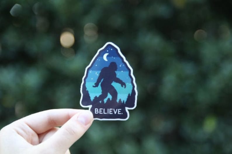 May include: A blue and green sticker featuring a silhouette of Bigfoot walking through a forest. The sticker includes a crescent moon and the text "BELIEVE" printed on it.