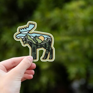 May include: A die-cut sticker featuring a moose silhouette filled with a landscape design. The design includes trees, mountains, a setting sun, and a blue sky. The sticker has a white border and is held against a blurred green background.