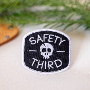 Beside a brass compass and a pine sprig, the Safety Third patch sits on a cream tabletop with leather nearby.