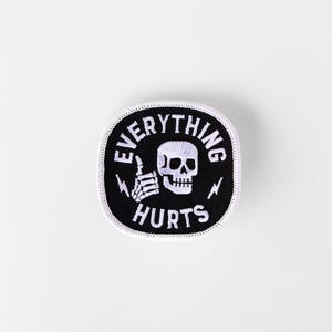 May include: Black and white embroidered patch with the text "EVERYTHING HURTS" surrounding a skull and a skeleton hand giving a thumbs up. Two lightning bolts flank the skull. The patch has a white border.