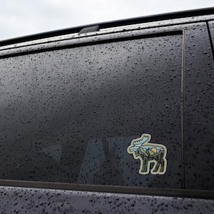 May include: A die-cut sticker featuring a stylized moose silhouette with a mountain and forest landscape design. The sticker has a light green outline, with blue, green, yellow, and brown details. The sticker is adhered to a dark, wet surface.