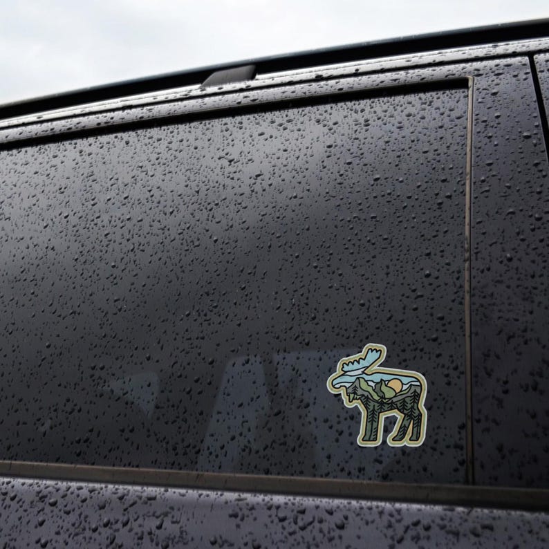 May include: A die-cut sticker featuring a stylized moose silhouette with a mountain and forest landscape design. The sticker has a light green outline, with blue, green, yellow, and brown details. The sticker is adhered to a dark, wet surface.