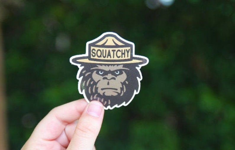 May include: A brown and black sticker of a cartoon Bigfoot wearing a ranger hat with the text "Squatchy" on it.