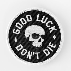 May include: A circular embroidered patch with a black background and a white border. The centre features a white skull graphic. The text GOOD LUCK is arched across the top and DON'T DIE is arched across the bottom in white lettering.