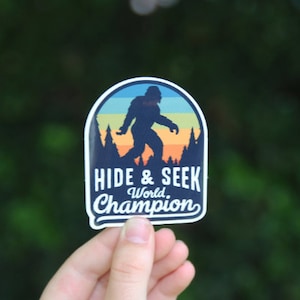 May include: A blue and orange sticker featuring a silhouette of Bigfoot walking through a forest. The text reads "Hide & Seek World Champion".
