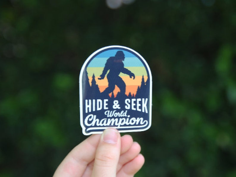 May include: A blue and orange sticker featuring a silhouette of Bigfoot walking through a forest. The text reads "Hide & Seek World Champion".
