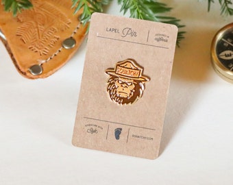 May include: A gold-colored lapel pin featuring a cartoon Sasquatch wearing a ranger hat with the word "SQUATCHY" printed on it. The pin is on a brown card with the text "LAPEL Pin" and "Adventure with Style" printed on it.